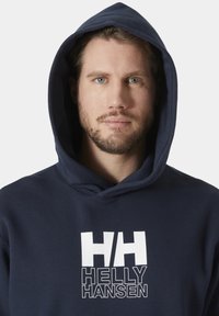 Marineblauwe hoodie met voorste kangoeroezak, groot wit logo "HH" en "HELLY HANSEN" bedrukt. Zachte stof, verstelbare trekkoordcapuchon.