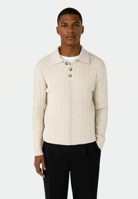 Kremfarget ribbestrikket genser med kragenek, tre knapper og en brodert midtpanel, brukt sammen med svarte bukser.