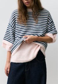 Kvinde iført lag-på-lag striktrøjer med lyserøde og marineblå striber, mørke jeans og sølvringe, løfter den yderste trøje for at afsløre den inderste.