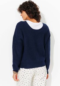 Pull en maille bleu marine avec un large décolleté et des épaules tombantes, associé à un pantalon blanc à motifs avec de petits motifs floraux bleus.