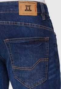 Dunkelblaue Jeans aus Denim mit oranger Naht. Die Merkmale umfassen eine Gesäßtasche und ein hellbraunes Lederetikett mit Logo und Text am Bund.