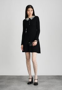 Vrouw in een zwarte jurk met een witte kanten kraag, gestippelde witte panty's, zwarte Mary Jane hakken en een kleine zwarte handtas in haar hand.