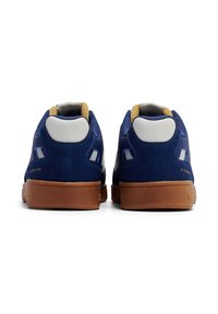 Blå mockaskor med vita läderdetaljer och brun gummisula. Har en vadderad krage och perforerade sidor för andningsförmåga.