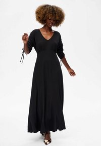 Femme aux cheveux bouclés portant une longue robe noire avec des manches froncées et des chaussures pointues à motifs noir et blanc, se tenant sur un fond blanc.