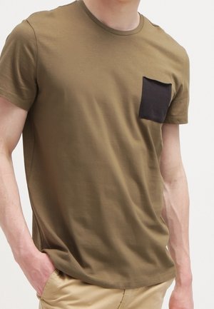 Homme portant un T-shirt à manches courtes vert olive avec une poche poitrine noire et un pantalon beige, debout avec une main dans la poche.