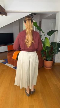 Γυναίκα με μακριά αλογοουρά, φορώντας μπορντό μπουφάν και μπεζ φούστα, στέκεται ξυπόλητη σε ξύλινο πάτωμα σε σαλονάκι με φυτά και τηλεόραση.