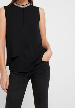 Černý top bez rukávů s vrstveným předním dílem a perlami na výstřihu, nošený s černými džínami s malými dekorativními cvočky na oblasti kapes.