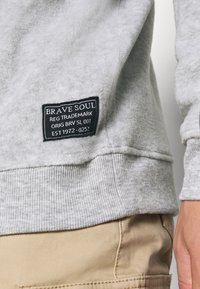 Grå fleece-tröja med en svart märkeetikett. Halsringningen och fållen har en ribbad struktur. Materialet verkar mjukt och varmt.