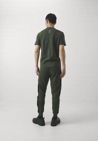 Pánský sportovní outfit zahrnuje přiléhavé zelené tričko a zúžené zelené kalhoty s černými postranními detaily, vyrobené z měkké, prodyšné látky.