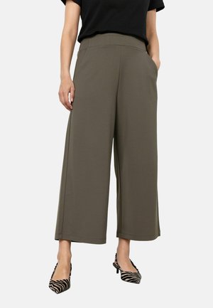 Femme portant un pantalon large court olive, un t-shirt noir et des escarpins à talons bas pointus imprimé zèbre, debout les mains dans les poches.