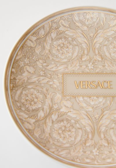 Okrągły talerz Versace z misternymi złotymi wzorami kwiatowymi na kremowym tle, z wyraźnym złotym logo "VERSACE" w centrum.