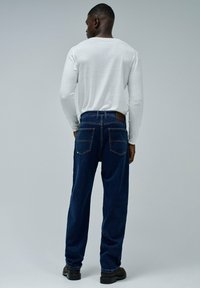 Vaqueros sueltos de color azul oscuro con dos bolsillos traseros, combinados con una camisa blanca de manga larga. Textura lisa y detalles mínimos visibles.