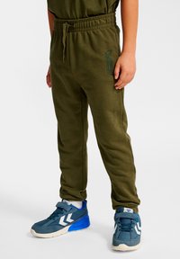 Pantalones de chándal verde oliva con cintura elástica y cordón. Combinados con zapatillas azules con detalles blancos y un diseño texturizado.