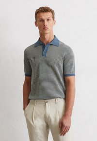 Polo w paski w niebiesko-beżowych kolorach, posiada jednolity niebieski kołnierzyk, krótkie rękawy i dopasowany krój. W komplecie z jasnobeżowymi spodniami.