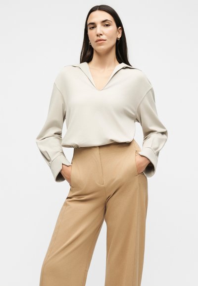 Γυναίκα με μακριά σκούρα μαλλιά, που φοράει κρεμ μακρυμάνικη μπλούζα με V-λαιμόκοψη και ψηλόμεσο καμηλό παντελόνι με φαρδιά πόδια, με τα χέρια στις τσέπες.