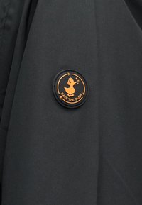 Μαύρο ύφασμα με στρογγυλό μαύρο και πορτοκαλί έμβλημα που απεικονίζει μια πάπια που τραγουδά και το κείμενο "SAVE THE DUCK" περιβαλλόμενο από κυκλικά βέλη.