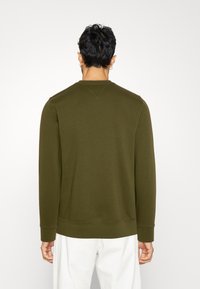 Felpa verde oliva con maniche lunghe, collo rotondo e polsini a costine. Tessuto morbido e cuciture sulle spalle. Indossata sopra pantaloni bianchi.