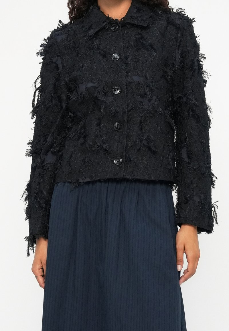 Femme portant une veste noire texturée boutonnée à manches longues sur une jupe plissée foncée.