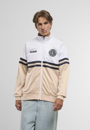 Young man wearing a white and beige zip-up jacket with black stripes and patches, paired with light blue jeans against a plain background.