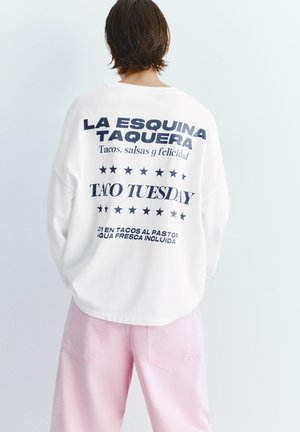 Person wearing white sweatshirt with black text about tacos and "Taco Tuesday," paired with light pink pants, standing against a plain white background.