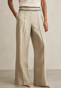 Beige wijde broek van linnen met verticale plooien en een zwarte gestreepte tailleband. Gecombineerd met puntige beige hakken.