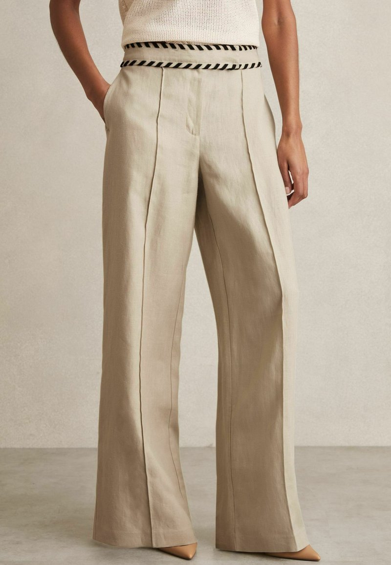 Beige wijde broek van linnen met verticale plooien en een zwarte gestreepte tailleband. Gecombineerd met puntige beige hakken.