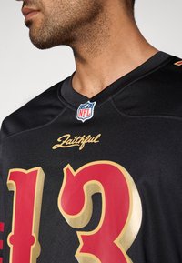 Μαύρη φανέλα NFL με μεγάλα κόκκινα και χρυσά νούμερα "13". Ο λαιμός φέρει στρογγυλό έμβλημα NFL και τη λέξη "Faithful" σε χρυσά γράμματα.
