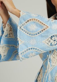 Hellbluse mit Glockenärmel, verziert mit filigraner weißer Stickerei, Spitzenbesatz und floralen Mustern auf dem gesamten Stoff.