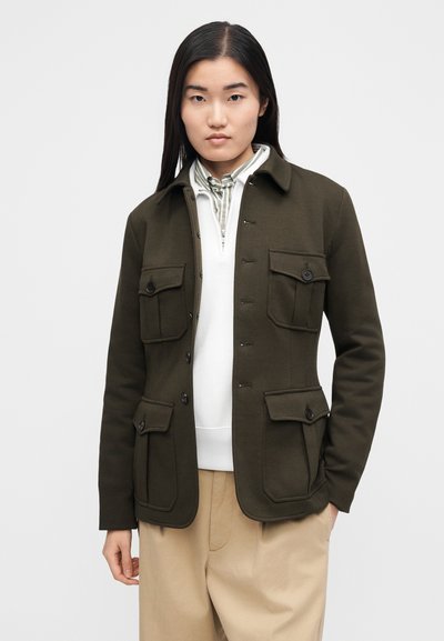 Maslinasto zelena jakna z gumbi spredaj, dvema velikima žepoma na prsih in mehko teksturo. Oblečena čez belo majico z manj kot polovico zadrge na sprednji strani.