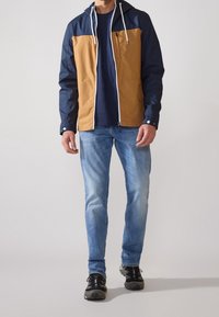 Man som bär en tvåfärgad marinblå och sandfärgad zip-up-jacka, marinblå skjorta, ljusblå jeans och svarta sneakers som står mot en enfärgad bakgrund.