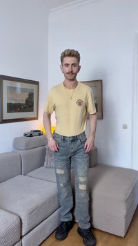 Camiseta de algodão amarela com um logo, jeans azuis desgastados com rasgos e sapatilhas pretas. De pé num espaço de estar com móveis cinzentos.