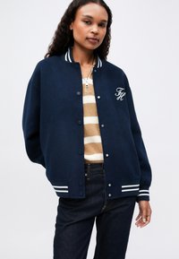 Žena oblečená v námorníckej varsity bunde s bielymi pruhmi a iniciálami "FH", skombinovaná s bežovým pruhovaným sveterom a tmavými džínsami.