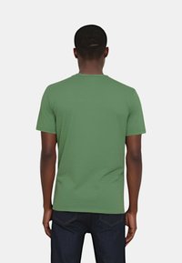 T-shirt verde a maniche corte realizzato in tessuto morbido, con scollatura rotonda e vestibilità morbida, visto di spalle. I jeans sono visibili in vita.