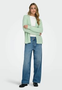 Light green knitted cardigan with long sleeves, open front, worn over a white top and loose blue jeans. Black ankle boots complete the outfit.