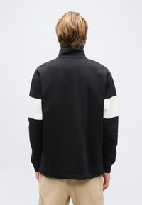 Άτομο με κοντά ανοιχτό καστανά μαλλιά, που φοράει μαύρο μακρυμάνικο πουκάμισο με λευκές λωρίδες στους πάνω βραχίονες, μπεζ παντελόνια, βλέποντας προς τα πίσω.