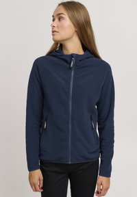 Felpa in pile blu navy con zip frontale completa, due tasche laterali con zip e cappuccio foderato. Caratterizzata da una superficie liscia e una leggera elasticità.