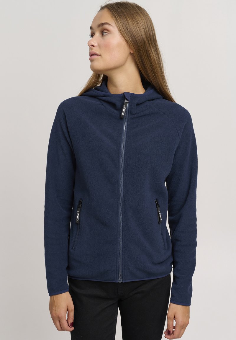 Felpa in pile blu navy con zip frontale completa, due tasche laterali con zip e cappuccio foderato. Caratterizzata da una superficie liscia e una leggera elasticità.