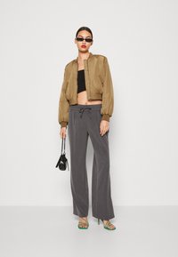 Veste bomber en satin marron, top noir court, pantalon large gris, sandales à talons vertes et petit sac à main noir avec chaîne.