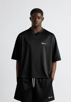 Jeune homme portant un t-shirt oversize en maille noire et un short assorti avec le logo « Buuoco », debout, les mains dans les poches, devant un fond uni.