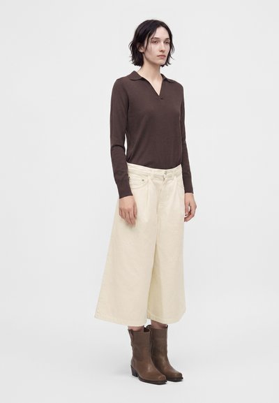 Femme aux cheveux courts et foncés portant un haut marron à manches longues, un pantalon large court crème et des bottines marron, debout devant un fond uni.