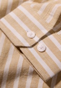 Gestreept shirt met afwisselende beige en witte kleuren. Heeft een kraag, twee witte knopen en zichtbare stiksels op een gestructureerde stof.