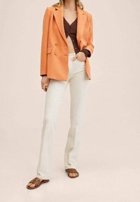 Veste blazer orange à double boutonnage, haut marron boutonné, jean blanc évasé et sandales marron à brides. Texture lisse, design ajusté.