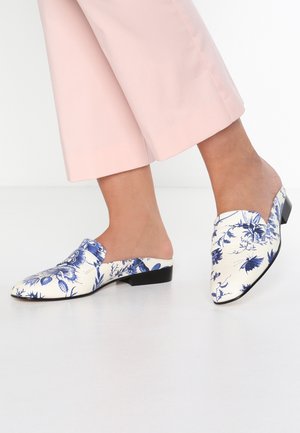 Pieds portant des chaussures blanches sans lacets avec des motifs floraux bleus, associées à un pantalon court rose clair sur un fond uni.