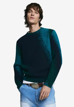 Joven con suéter de textura verde azulado oscuro, jeans azul claro, hebilla de cinturón con diseño y pendiente de aro plateado, posando frente a un fondo blanco.