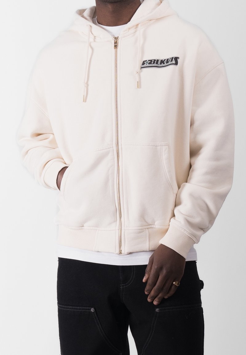 Homme portant un sweat à capuche crème zippé avec poches avant et logo noir sur la poitrine, associé à un pantalon noir et un sous-vêtement blanc.