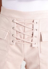 Pantalon rose clair en tissu doux et texturé avec un laçage croisé à l'avant et deux poches latérales à rabat.