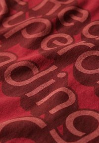 Czerwona tkanina z wzorem dużych, nachodzących na siebie liter w różnych odcieniach czerwieni i brązu, tworząca teksturowany wygląd.