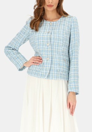 Kobieta ubrana w jasnoniebieską i białą kratowaną kurtkę z fakturą i srebrnymi guzikami, w połączeniu z białą plisowaną spódnicą, pozująca z ręką na biodrze.