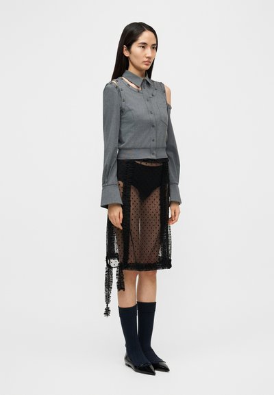 Γκρι πουκάμισο με κουμπιά και σχεδιασμό με αποκαλυπτούς ώμους, σε συνδυασμό με μαύρη διαφανή φούστα με πουά. Μαύρες κάλτσες μέχρι το γόνατο και μυτερές μαύρες γόβες.