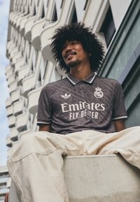 Grå polo shirt med struktureret design, hvidt logo og teksten "Emirates Fly Better", parret med lyse bukser, sat mod moderne arkitektur.